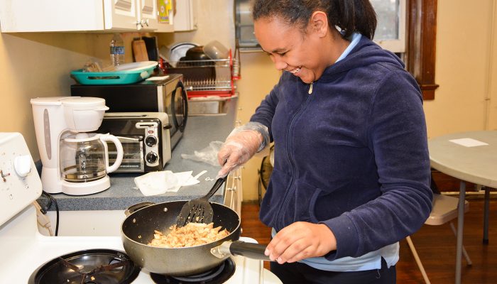 Jovem mulher, sorrindo enquanto cozinha algo em uma frigideira no fogão.