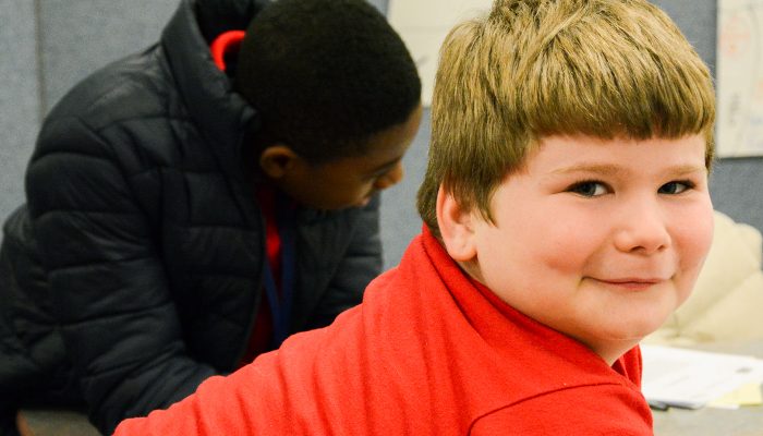 A white boy in a red shirt leans forward and smiles slightly.