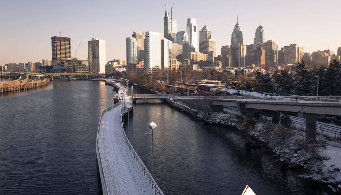 Skyscrapers rise far off in the distance. They are tall and thin, many of them built of glass. The Schuylkill River winds toward the buildings, and the Schuylkill River Walk provides walkers and bicyclists a space to travel up the river. There is a light dusting of snow covering everything. Nobody is around. It is very cold but the sky is clear and the sun is out.