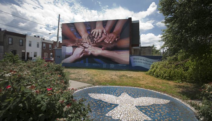 A mural depicts multicultural hands overlapping one another communicating peace, teamwork, and cooperation. The mural is on the side of a rowhouse in South Philadelphia. There is a community green space in front of it with bushes and a mosaic on a patio depicting a dove.