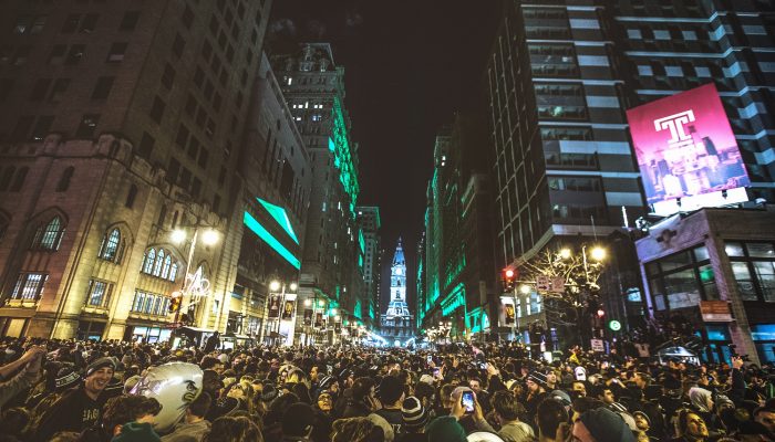 Thousands upon thousands of people are smiling, screaming happily, and waving Eagles flags on Broad Street. It is nighttime, and everyone ran outside to celebrate after the Eagles won the Super Bowl. The crowd is massive. City Hall is in the far distance.