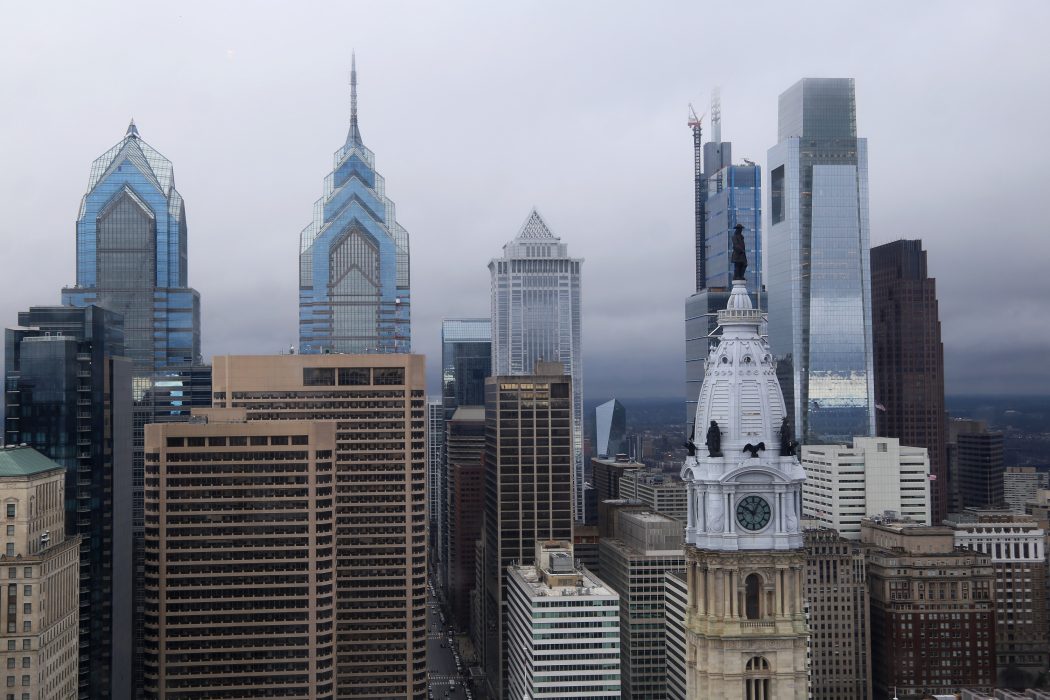 The skyline of Philadelphia shows tall buildings rising high above the street. The City Hall clock tower is on the foreground with the statue of William Penn atop it. It is a wet, cloudy, cold day. The new Comcast tower is almost finished in the background with a construction crane.