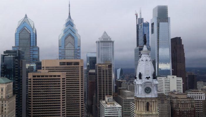 The skyline of Philadelphia shows tall buildings rising high above the street. The City Hall clock tower is on the foreground with the statue of William Penn atop it. It is a wet, cloudy, cold day. The new Comcast tower is almost finished in the background with a construction crane.