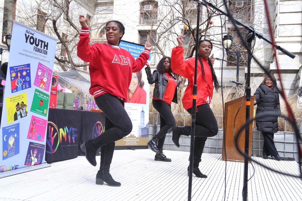 Two women from a sorority dance on a stage in City Hall's Courtyard while Councilwoman Blondell Reynolds Brown looks on. There are banners behind them promoting Women's History Month events. Everyone looks very happy.