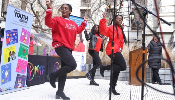 Two women from a sorority dance on a stage in City Hall's Courtyard while Councilwoman Blondell Reynolds Brown looks on. There are banners behind them promoting Women's History Month events. Everyone looks very happy.
