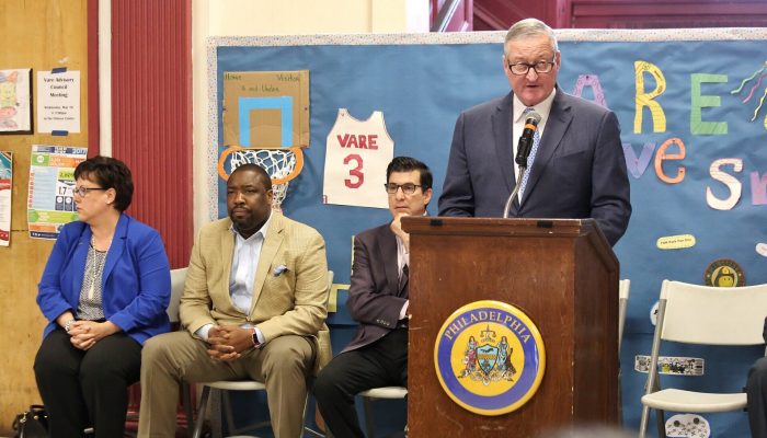 Mayor Kenney stands at a podium while Councilman Johnson sits next to him. There is art showing a basektball, hoop, and sports jersey behind them. The art is made out of construction paper and seems to have been created by children.