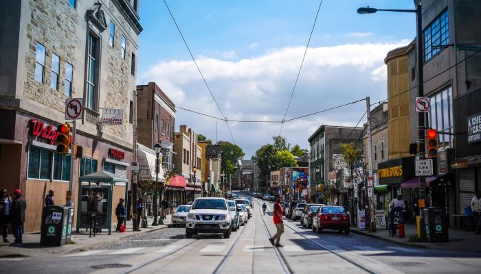 A busy street in Philadelphia has trolley tracks in the middle of it. There are businesses everywhere and cars and people moving around. It's a very active commercial corridor with a variety of businesses and shops, some chains and many local and independent.