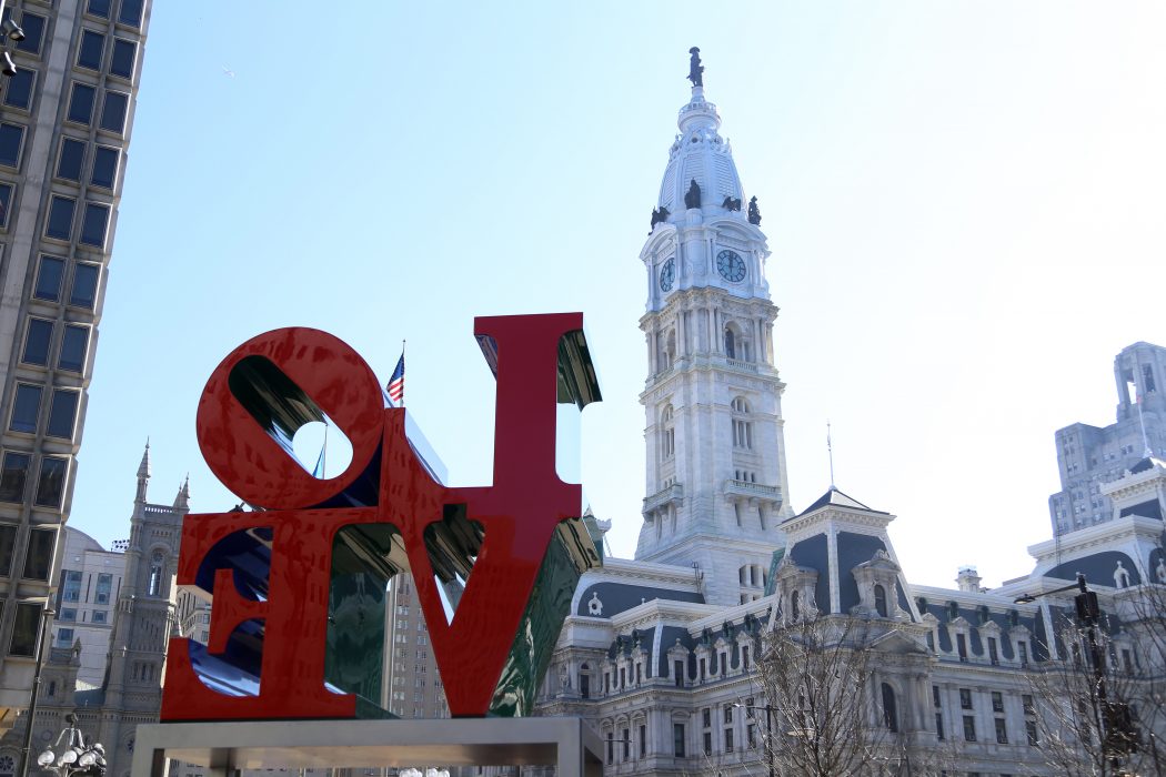 City Hall appears behind the LOVE Statue in JFK Plaza, otherwise known as LOVE Park. The clock tower with the William Penn Statue rises in the distance. City Hall is very large compared to the statue. It is a sunny day.