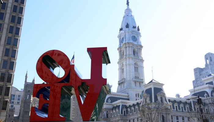 City Hall appears behind the LOVE Statue in JFK Plaza, otherwise known as LOVE Park. The clock tower with the William Penn Statue rises in the distance. City Hall is very large compared to the statue. It is a sunny day.