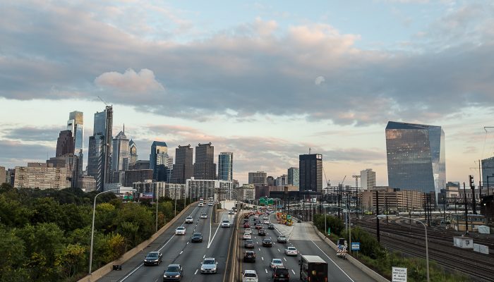 The Schuylkill Expressway is filled with cars driving in and out of Center City Philadelphia. Glass and steel buildings rise from the horizon and the trees of Fairmount Park are to the side in the foreground.