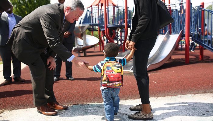 Mayor Kenney bends down to shake the hand of a little boy on a playground in Philadelphia. There are slides and a jungle gym nearby. The little boy is about three or four years old. The Mayor is smiling and happy.