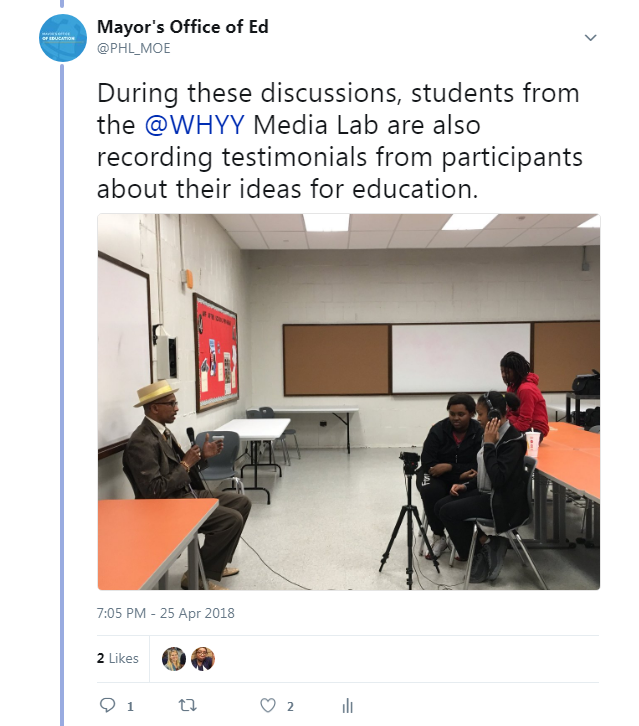 A man with a hat on speaks into a microphone as students sit by and record his testimonial to the school board 