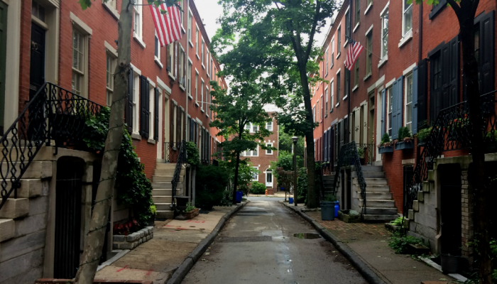 A narrow, residential street in Philadelphia features row homes, trees and American flags