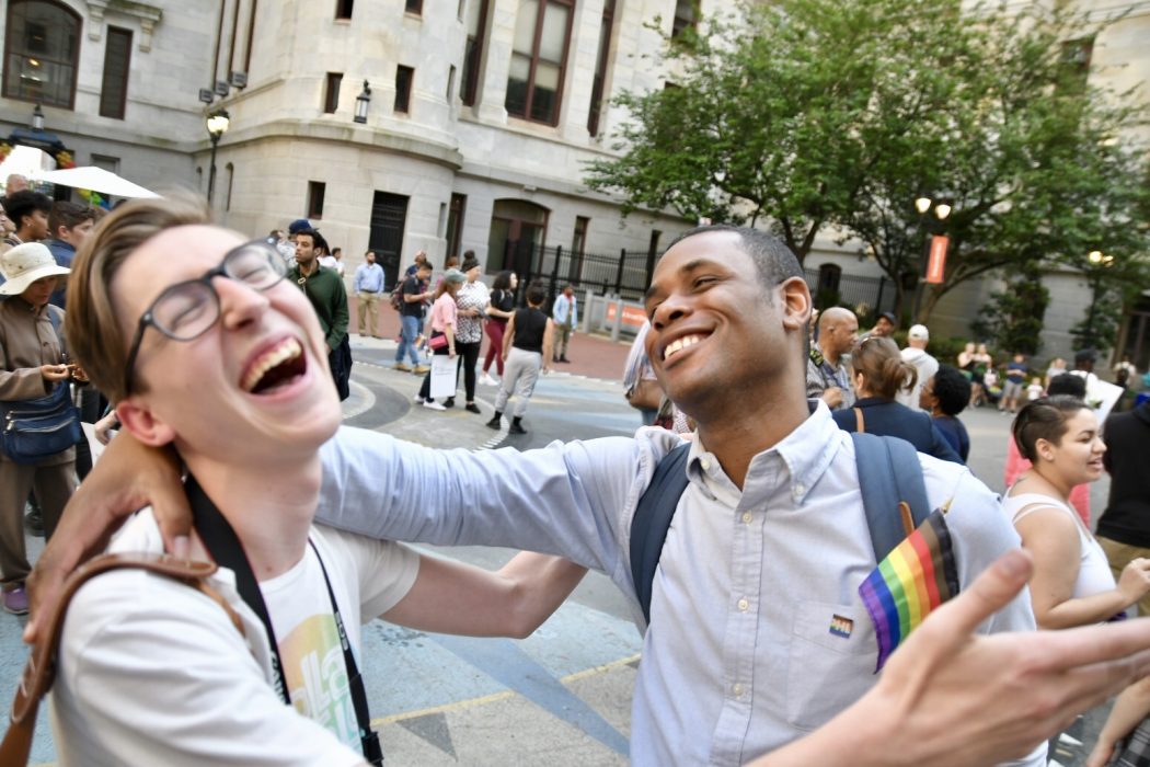 two people hugging and laughing at city hall's courtyard with a rainbow pride flag during a pride month event.