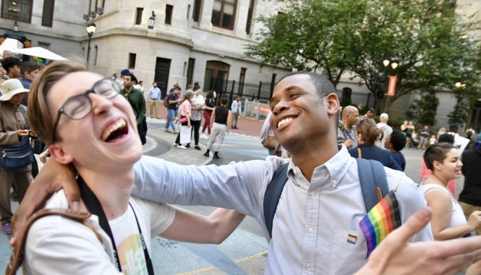 two people hugging and laughing at city hall's courtyard with a rainbow pride flag during a pride month event.