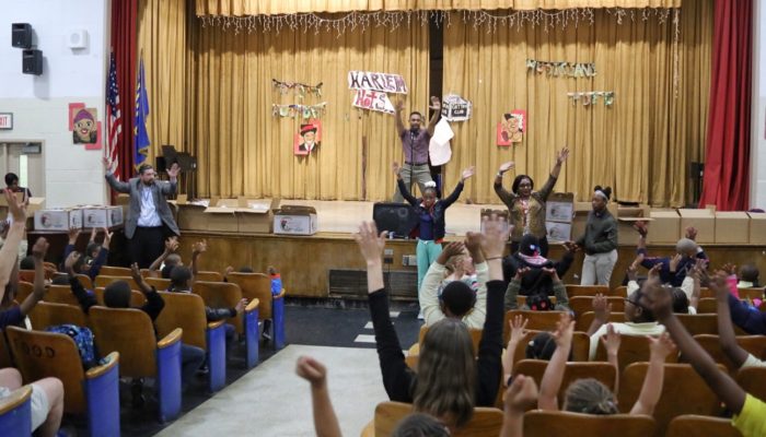 Gregory Wright stands on stage at Gideon with his hands in the air, students in the auditorium follow along, and do the same