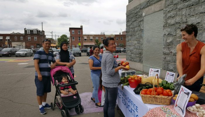 Community members stand at a farmstand with fruit in hand, Beth Dougherty smiles behind the table