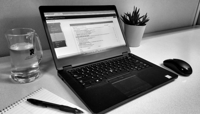 A laptop sits on a desk surrounded by a mug, a small plant, a computer mouse and a notepad