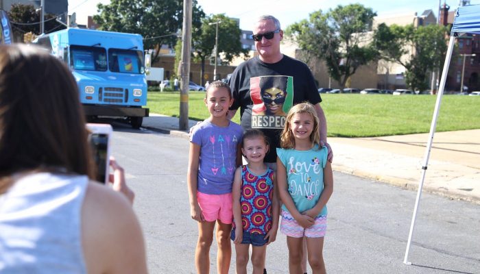 Mayor Kenney wears an Aretha Franklin t-shirt that says, "RESPECT," as he stands next to three girls at the PHL Unity Cup Block Party; there is a food truck behind them and a soccer field nearby.