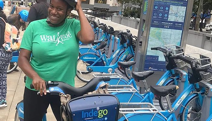 A smiling woman on a bike stands next to a bike share rack.
