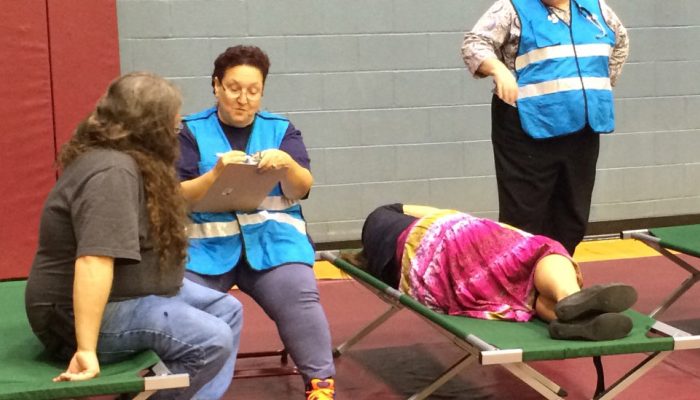 People in blue vests talk to people on cots.