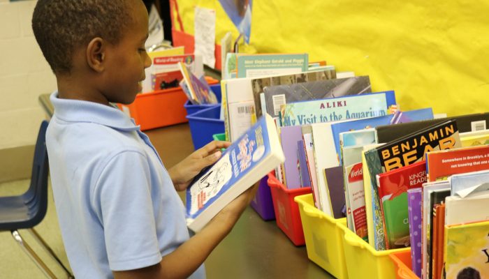 A young student browses a table full of books, picks one up, and reads through it, smiling