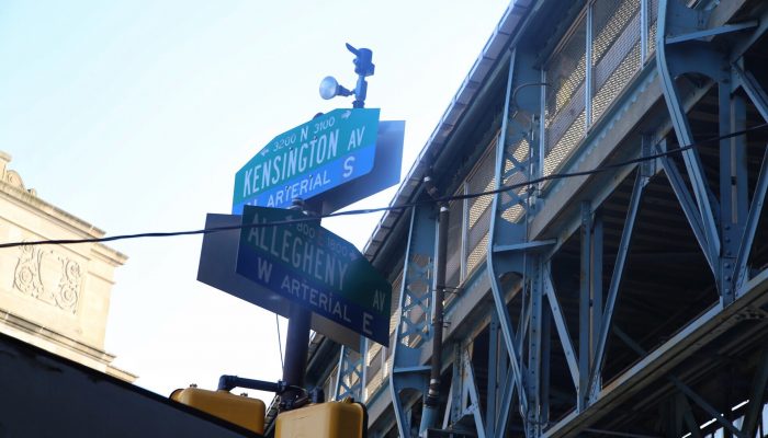 Street signs showing the intersection of Kensington and Allegheny avenues with the Market-Frankford elevated train track or "El" in the background.