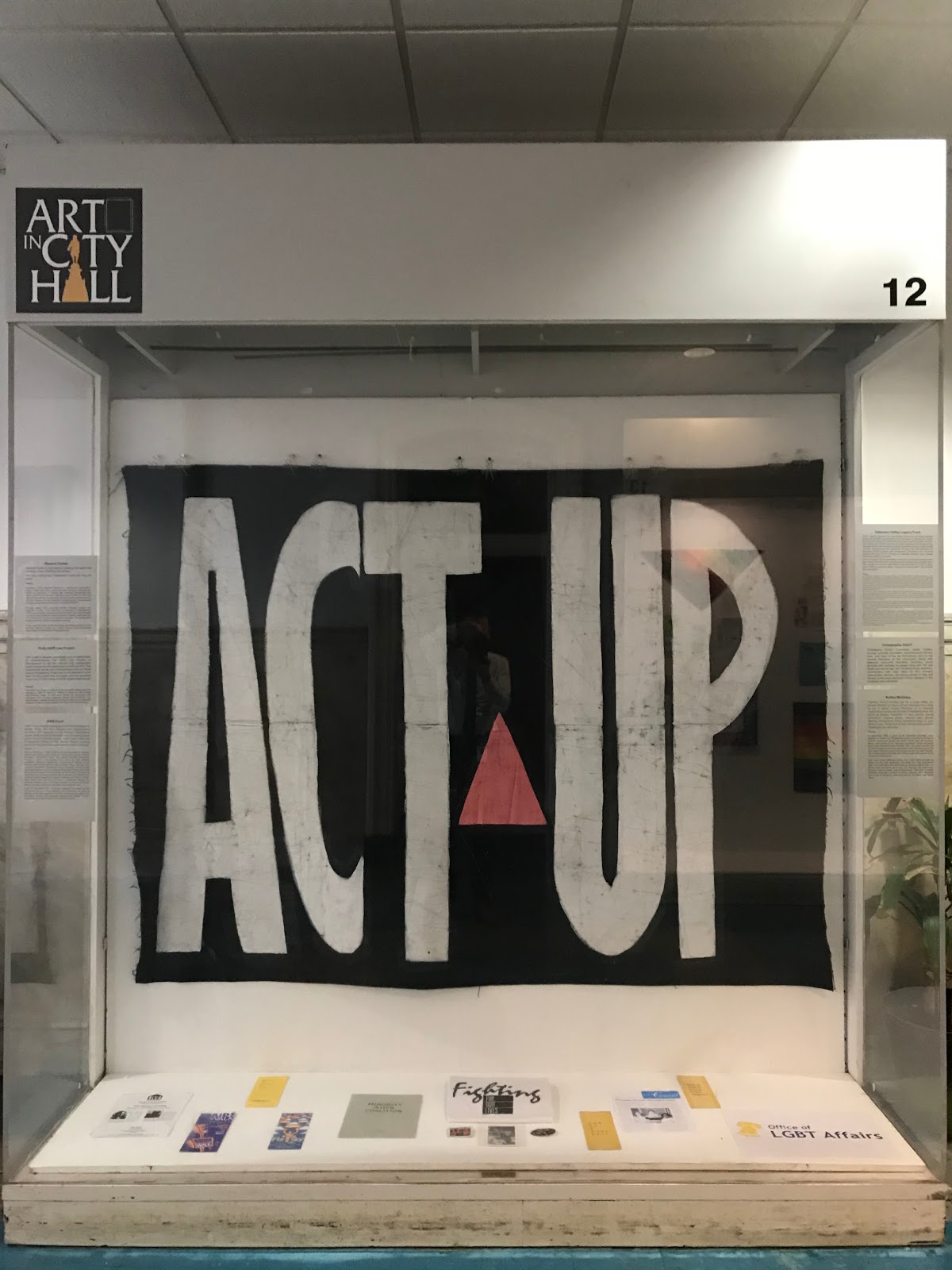 art exhibit in city hall showing different lgbtq artifacts including a large "act up" banner