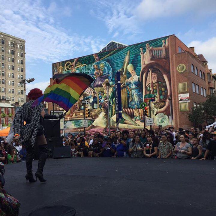 people gather in front of a large mural outside with the more color more pride flag waving in the wind