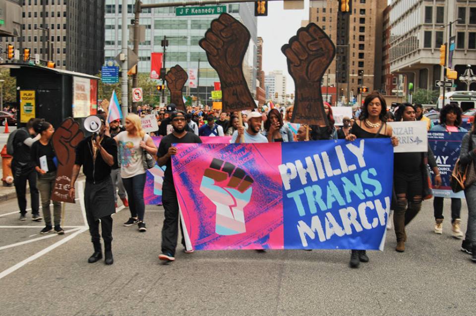 people marching down the street with a big banner that says philly trans march