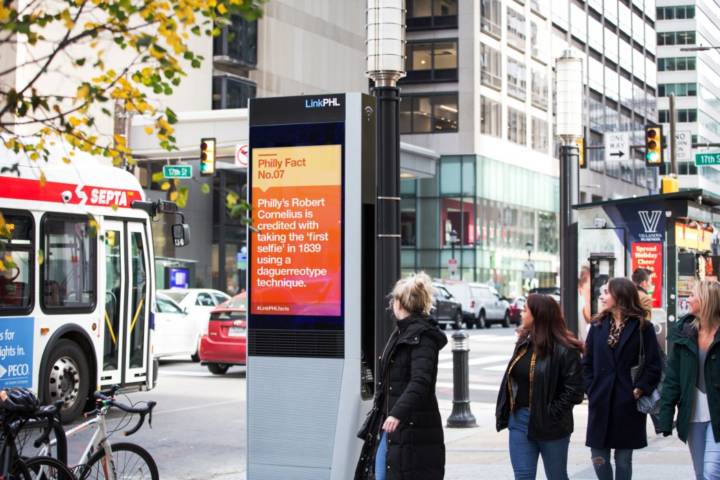 A SEPTA bus driving by a new LinkPHL kiosk as passersby smile and look at the new kiosk.