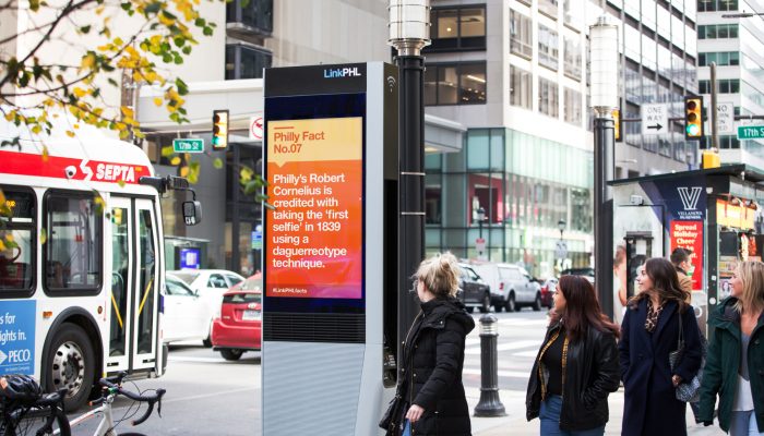 A SEPTA bus driving by a new LinkPHL kiosk as passersby smile and look at the new kiosk.