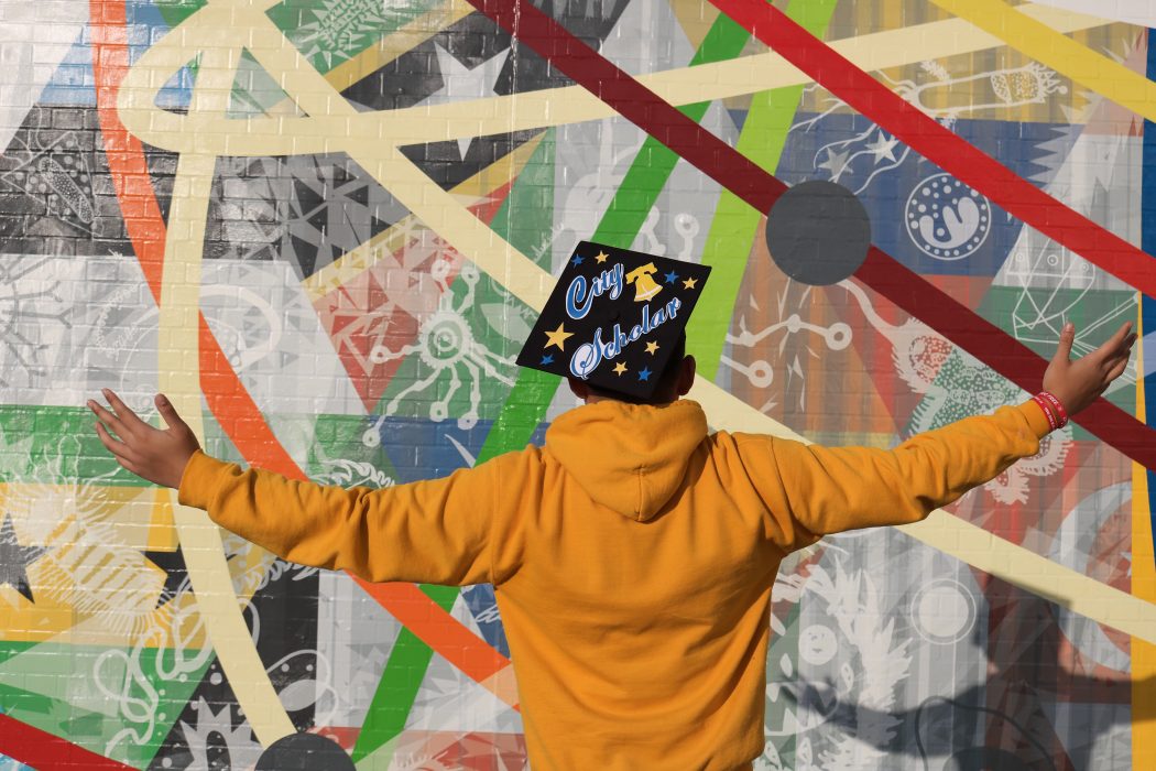 A student stands in front of a mural at South Philadelphia High School with a graduation cap that reads "City Scholar"