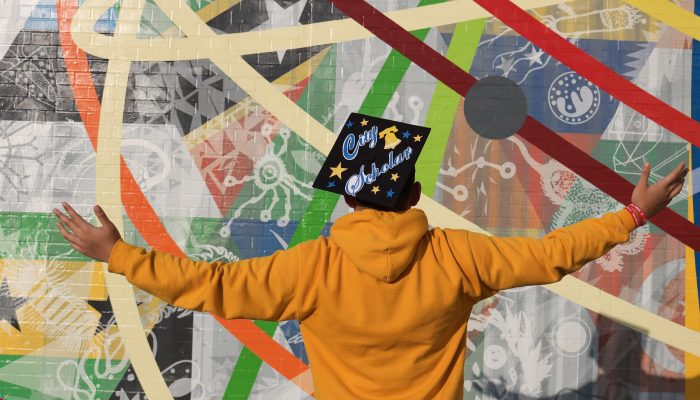 A student stands in front of a mural at South Philadelphia High School with a graduation cap that reads "City Scholar"