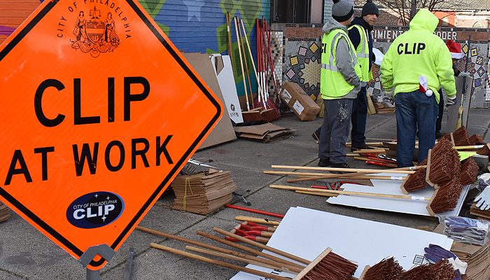 An orange sign says 'CLIP at work' and three workers stand next to brooms.