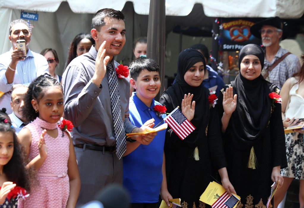 Children and youth of various ages holding their hands up to swear their oaths as new U.S. citizens during a naturalization ceremony at City Hall during the July 4th festivities; a boy is holding an American flag.