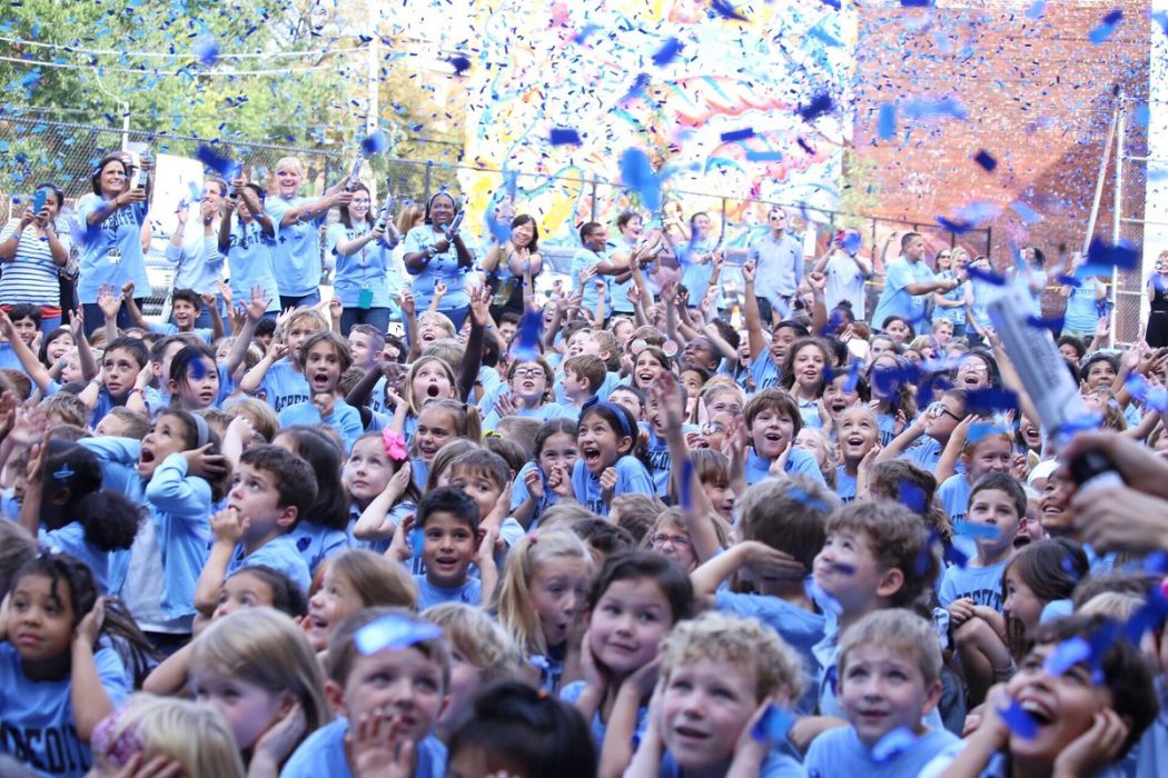 Kids cheering happily after their school, Meredith, was named a national blue ribbon school this year; there are thousands of pieces of blue paper falling from the sky in a ticker-tape parade style celebration.