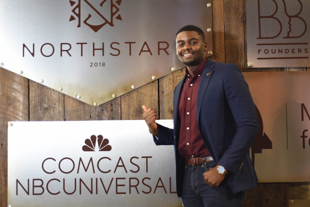 An attendee uses his thumb to point to a Project Northstar sign above a Comcast/NBC Universal sign at the tech conference for Black and Brown startup leaders and professionals. 