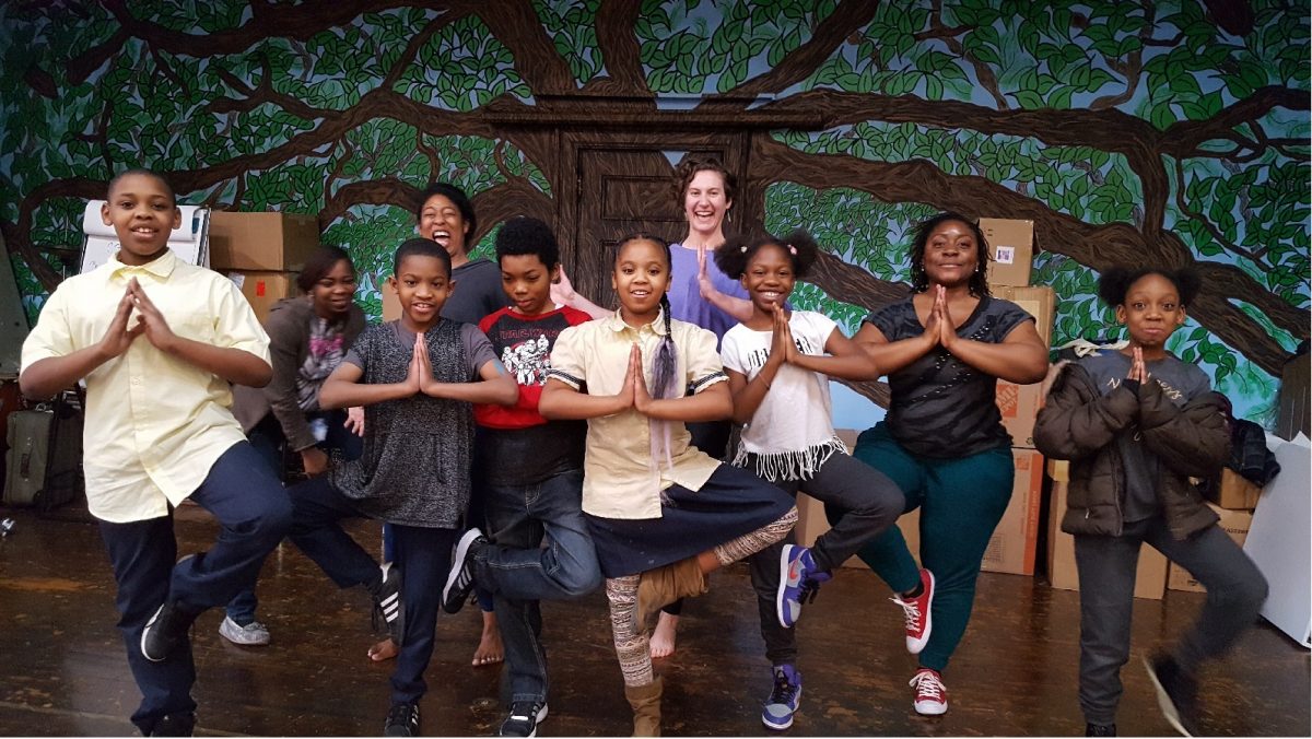 Students and Laura Crandall, Community Schools' Healthy Schools Specialist stand in a yoga pose with one leg raised and arms together smiling