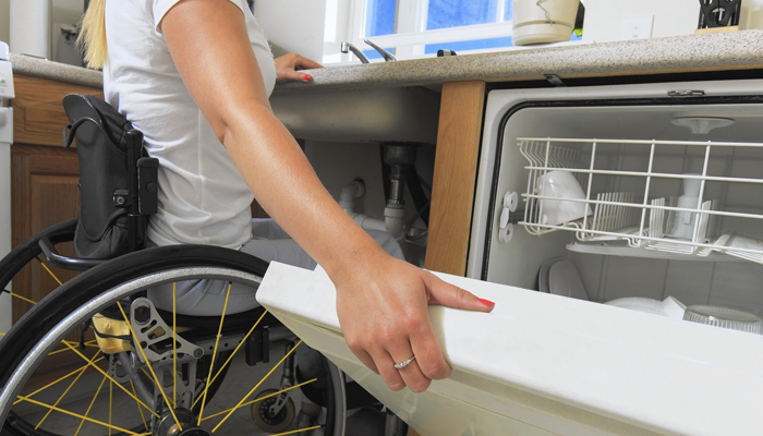 A woman in a wheelchair closes a dishwasher.