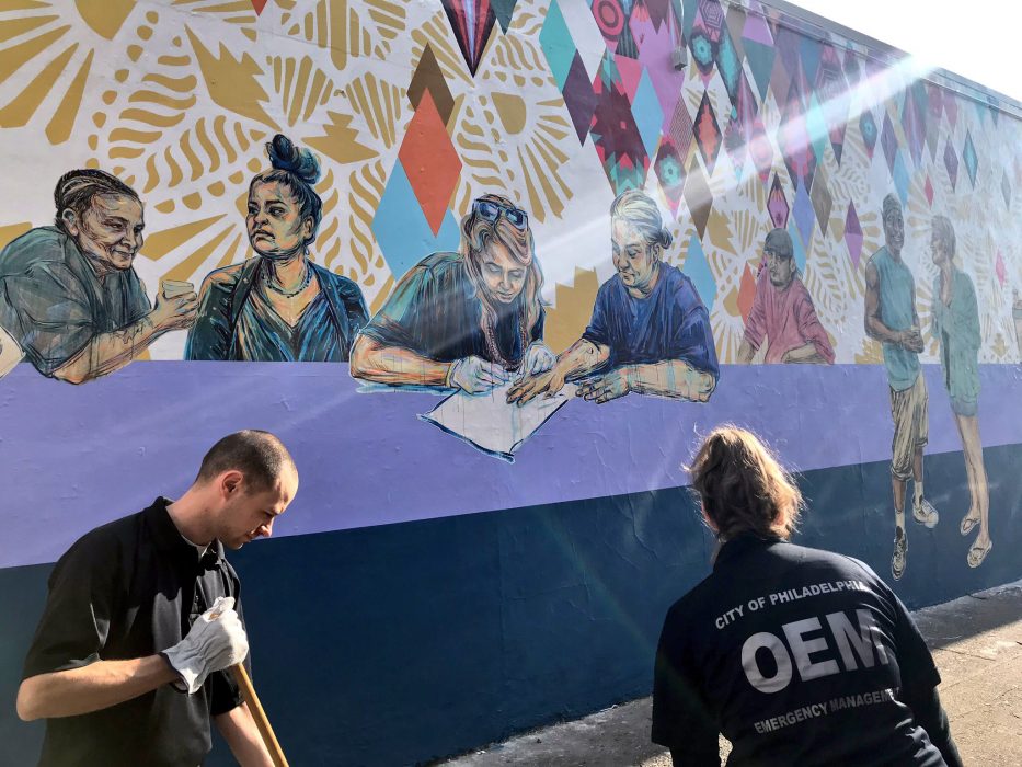 Two people sweeping the sidewalk in front of a mural.