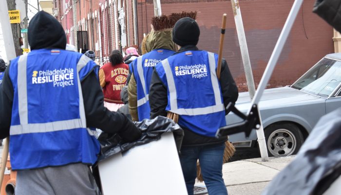 Group of people wearing Philadelphia Resilience Project vests walk down a street carrying trashcans and cleaning equipment.