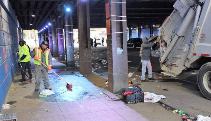 At a just-cleared encampment site, people clean up trash, blankets, and other supplies under a bridge.