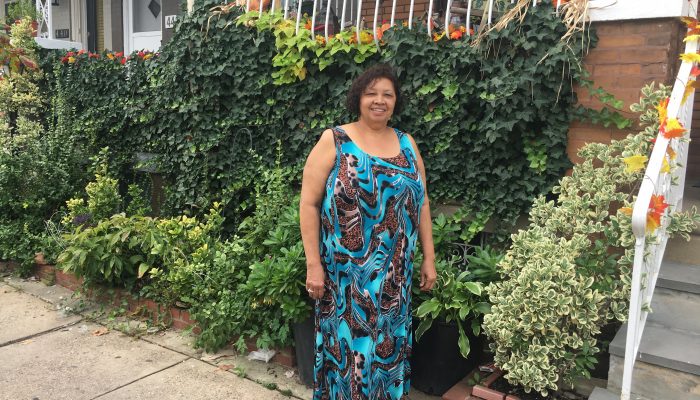 Catalina Hunter, a resident of Hunting Park, stands outside of her home. She is wearing a long, patterned blue and brown dress. She stands in front of a lush front yard garden