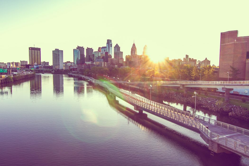 Philadelphia skyline. Sun is shining and creates a slight glare that peeks out behind the Philly skyline and over the bridges and water.