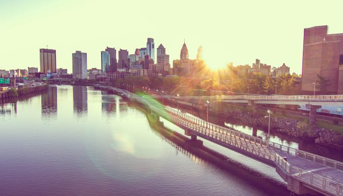 Philadelphia skyline. Sun is shining and creates a slight glare that peeks out behind the Philly skyline and over the bridges and water.