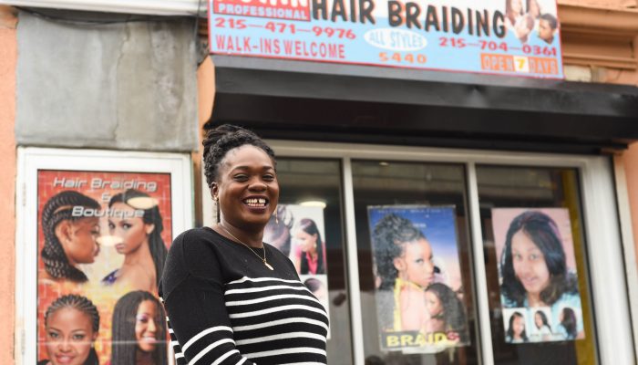 A black woman named Astan Sacko stands smiling in front of her hair braiding business in West Philadelphia.