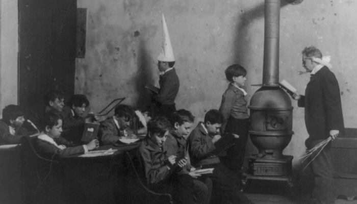 An old photograph shows a schoolroom with a teacher and students, one wearing a dunce cap