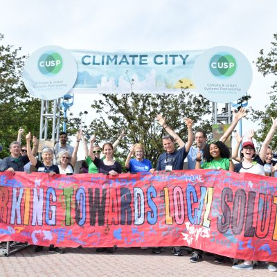 A group of people pose behind a large, colorful banner which reads: "working towards local solutions." Many are smiling and have their hands raised in the hair. The background is bright but cloudy. There are trees and another large banner which reads: "CUSP Climate City."