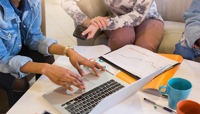 Uma mulher usa um laptop em uma mesa de café com outras pessoas reunidas ao seu redor.